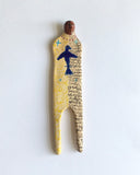 Elham Hemmat's 'Docile Bodies 7' a ceramic wall sculpture, measuring 17cm x 4cm x 1cm. Artwork contains colours of yellow and blue. Photographed mounted on the wall.