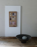 Abstract art piece on wood, on a white wall with a black bowl containing a grey pebble below.