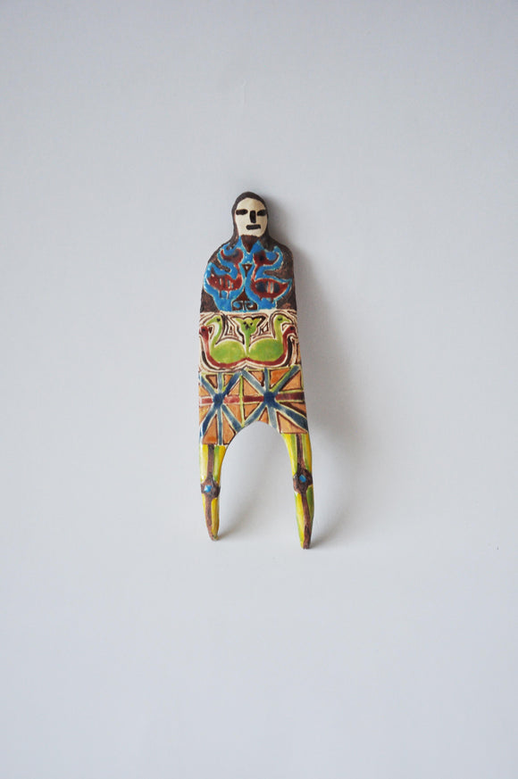 Elham Hemmat's 'Docile Bodies 4' a ceramic wall sculpture, measuring 14 cm x 6 cm x 1 cm. Artwork contains colours of green, yellow, blue, red and orange. Photographed mounted on the wall